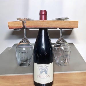 Peut inclure: Un porte-bouteille en bois avec deux verres à vin suspendus. Une bouteille de vin rouge est au centre du porte-bouteille. L'étiquette de la bouteille indique "Vieilles Vignes, Vins rouges de la Loire, Chinon".