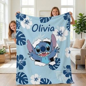 May include: A light blue blanket featuring the character Stitch bursting through a torn hole. The blanket is decorated with white and blue hibiscus flowers and the name "Olivia" in dark blue lettering. The blanket is held up by two people.