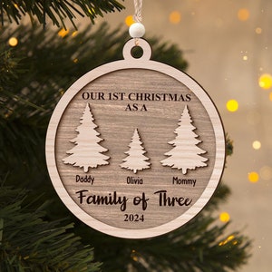 Könnte beinhalten: Ein runder Weihnachtsbaumschmuck aus Holz mit drei lasergeschnittenen Tannenbaumsilhouetten. Der Text lautet: OUR 1ST CHRISTMAS AS A, Daddy, Olivia, Mommy, Family of Three und 2024. Er enthält eine weiße Perle und eine Aufhängeschnur.