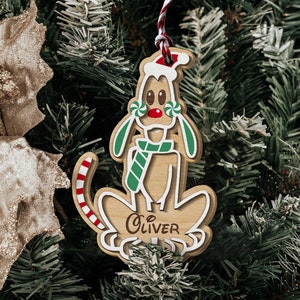 May include: A wooden dog-shaped Christmas ornament featuring a red and white Santa hat, a green and white striped scarf, and two green and white peppermint candies near the ears. The tail is red and white striped, and the name Oliver is engraved.