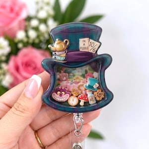 May include: A top hat shaped badge reel featuring a teal and purple plaid pattern. The hat displays a gold teapot, stacked teacups, and a sign reading IN A PUDDLE 10/6. Inside the clear center are miniature charms of a cat, clock, mushroom, bottle, and cookie.