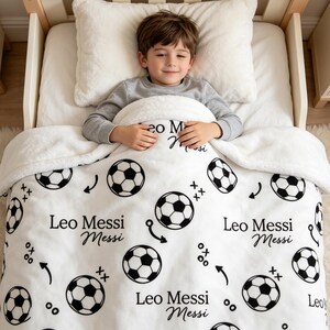 Op de afbeelding: Een witte pluche deken met een herhalend zwart patroon van voetballen, pijlen en de tekst Leo Messi. De deken ligt over een kind in een houten bed met een wit kussen en toont een zacht, gestructureerd ontwerp van de stof.