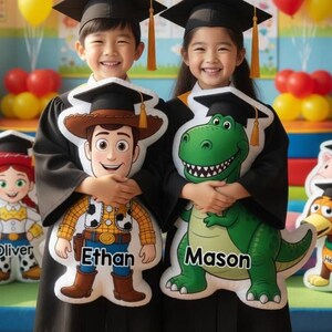 May include: Two children in graduation gowns holding character pillows, with the text "Congratulations Kindergarten Graduates!" Balloons and classroom decorations are in the background. The pillows feature Woody and Rex characters. Other character pillows include Buzz Lightyear, Jessie, and Slinky Dog.