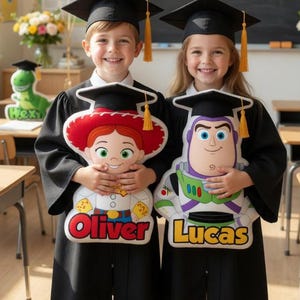 May include: Two children in graduation gowns holding character pillows, with the names "Oliver" and "Lucas" on them. The background includes a chalkboard that reads "CONGRATULATIONS CLASS OF 2026" and balloons.