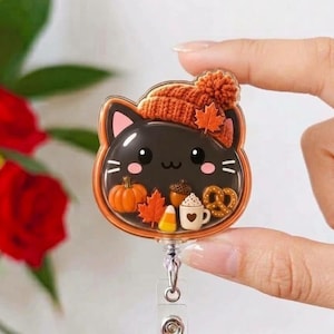 May include: A black cat-shaped badge reel featuring a textured orange knit hat with a maple leaf accent. The clear casing contains miniature autumn-themed charms including a pumpkin, maple leaf, acorn, candy corn, pretzel, and a coffee mug.