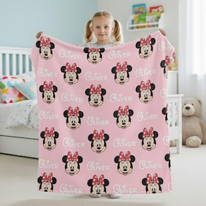 May include: A pink blanket featuring a repeating pattern of Minnie Mouse faces with red bows and the name "Oliver" in white script. The blanket is held up, showcasing its design and color.