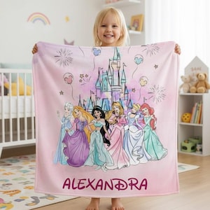 May include: A pink blanket featuring a castle and several cartoon princesses. The blanket has the name "ALEXANDRA" printed in a dark pink script. The princesses are wearing colorful gowns. The background has fireworks and balloons.