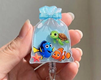 May include: A clear acrylic badge reel featuring a printed design of a plastic bag tied with a light blue bow. Inside the bag are illustrations of a blue tang fish, an orange clownfish, a green sea turtle, and a small pink starfish.