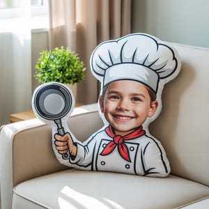 Puede incluir: Un cojín decorativo con la forma de un niño sonriente vestido de chef, sosteniendo una sartén. El cojín presenta un gorro de chef blanco, uniforme y un pañuelo rojo. El cojín está sobre un sofá beige.