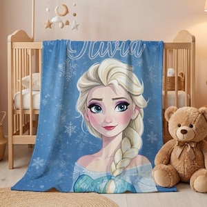 Pode incluir: Um cobertor de l&atilde; azul com uma ilustra&ccedil;&atilde;o de desenho animado de uma princesa, cabelo loiro e olhos azuis. O cobertor tem o nome "Olivia" em escrita branca e detalhes de floco de neve. Est&aacute; sobre um ber&ccedil;o.