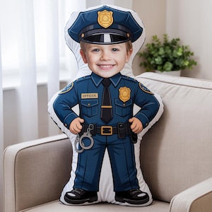 May include: A novelty pillow shaped like a police officer, featuring a child's face. The pillow is in a blue uniform with a badge, tie, and handcuffs. The child is smiling. The pillow is on a beige couch.