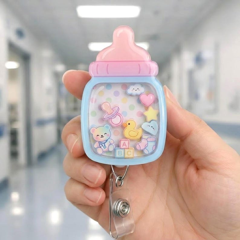 May include: A baby bottle-shaped badge reel with a light blue frame and a pink cap. The center features a shaker design with floating charms including a teddy bear, yellow duck, pacifier, heart, star, cloud, and an ABC block.