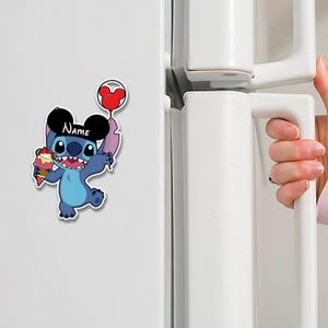 May include: A blue cartoon character magnet holding an ice cream cone and a heart-shaped balloon. The character has large ears and the word "Name" is written on its head. The magnet is attached to a white refrigerator.