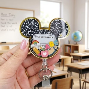 May include: A Mickey Mouse-shaped badge reel featuring a black and white speckled composition book pattern. Inside the clear casing are miniature charms including a rainbow, a yellow pencil, a pink apple with a heart, an open book, and the letters A, B, and C.