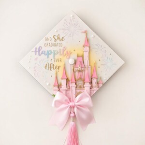 May include: A square graduation cap topper featuring a pink castle illustration, white firework accents, and the text "AND SHE GRADUATED Happily EVER After". The design includes small white pearls and a large, light pink satin bow with a matching tassel.