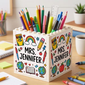 May include: A white block-style pencil holder decorated with school-themed illustrations, including rainbows, pencils, and globes. The text "MRS. JENNIFER" is prominently displayed. The holder is filled with various colored pencils, pens, and a ruler.