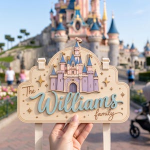 May include: A light-colored wooden sign featuring a layered cutout of a castle with blue and purple towers. The sign displays the text "The Williams family" in a blue script font, accented by small star shapes, and includes two fabric straps.