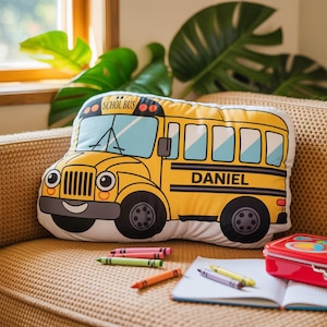 May include: A yellow school bus shaped pillow with the name "DANIEL" printed on the side. The pillow has black wheels, a smiling face, and the words "SCHOL BUS" on top. Crayons and a notebook are next to the pillow.