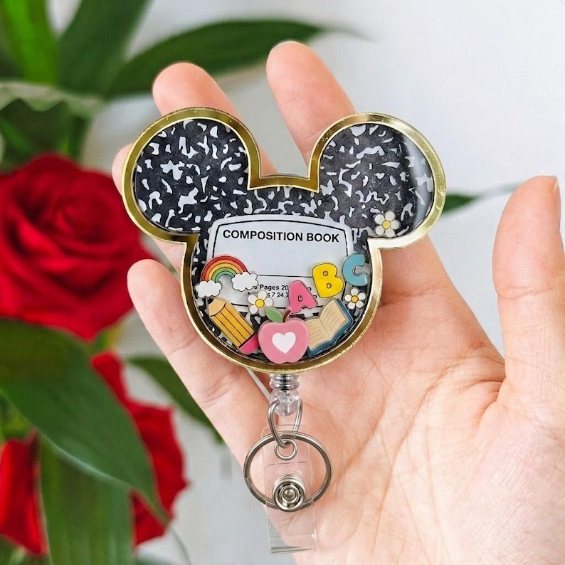 May include: A Mickey Mouse-shaped badge reel featuring a black and white composition book pattern. Inside the clear casing are miniature charms including a rainbow, a pencil, a pink apple, an open book, and the letters A, B, and C.