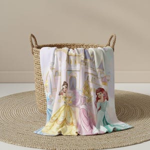 K&ouml;nnte beinhalten: Eine weiche, hellfarbene Decke mit Aquarellillustrationen von Prinzessinnen vor einem Schloss. Die Decke ist &uuml;ber einem geflochtenen Korb drapiert, darunter liegt eine runde, geflochtene Matte.