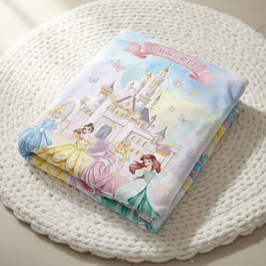 K&ouml;nnte beinhalten: Gefaltete Decke mit einem M&auml;rchenschloss und mehreren Prinzessinnen in farbenfrohen Kleidern. Die Decke hat einen hellblauen Hintergrund mit einem rosa Banner mit der Aufschrift "CHARLOTTE". Schmetterlinge und Sterne sind ebenfalls im Design enthalten.