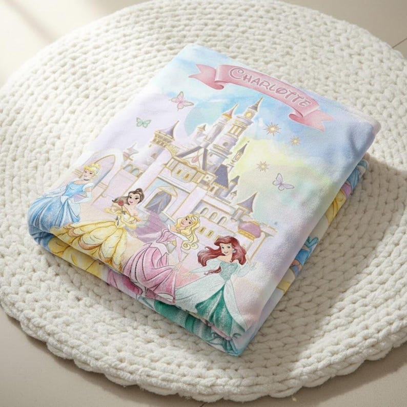 K&ouml;nnte beinhalten: Gefaltete Decke mit einem M&auml;rchenschloss und mehreren Prinzessinnen in farbenfrohen Kleidern. Die Decke hat einen hellblauen Hintergrund mit einem rosa Banner mit der Aufschrift "CHARLOTTE". Schmetterlinge und Sterne sind ebenfalls im Design enthalten.