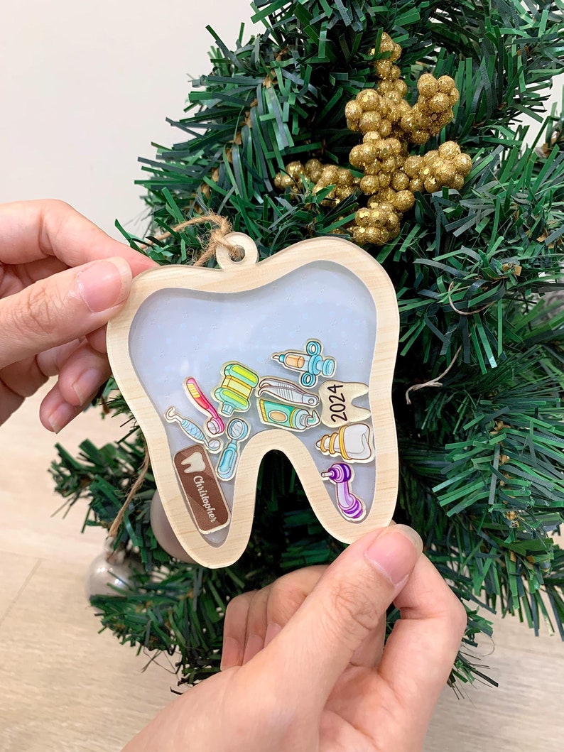 May include: A wooden-framed tooth-shaped Christmas ornament. Inside the clear center are miniature dental tools and a tooth graphic. The year "2024" is printed on the ornament, along with the name "Christopher". The ornament is hanging on a Christmas tree.