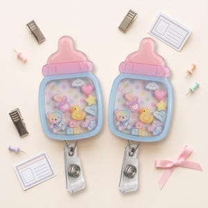 May include: Two baby bottle-shaped badge reels with a light blue frame and pink cap. The clear center contains floating charms including a bear, duck, pacifier, heart, star, cloud, and ABC blocks over a pastel polka dot background.