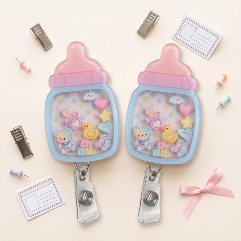 May include: Two baby bottle-shaped badge reels with a light blue frame and pink cap. The clear center contains floating charms including a bear, duck, pacifier, heart, star, cloud, and ABC blocks over a pastel polka dot background.