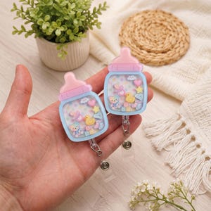 May include: Two baby bottle-shaped badge reels featuring a light blue base and a pink nipple top. The clear center contains floating miniature charms including a yellow duck, a teddy bear, a pink pacifier, a star, a heart, and alphabet blocks.