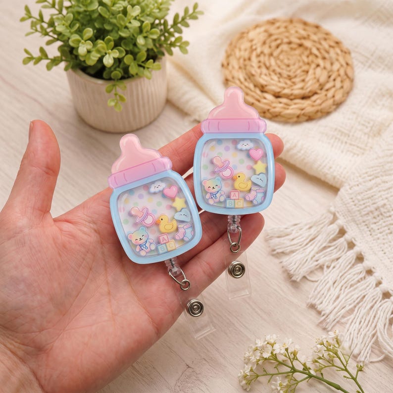 May include: Two baby bottle-shaped badge reels featuring a light blue base and a pink nipple top. The clear center contains floating miniature charms including a yellow duck, a teddy bear, a pink pacifier, a star, a heart, and alphabet blocks.