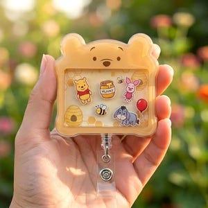 May include: A light yellow, bear-shaped badge reel featuring a clear window filled with small character icons including a yellow bear, a pink piglet, a gray donkey, a honey pot labeled HUNNY, bees, a beehive, and a red balloon on a white background.