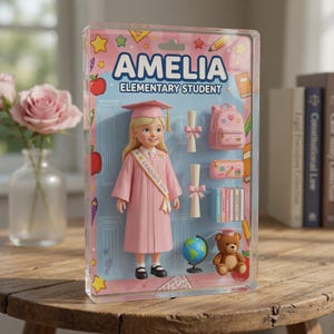 May include: A doll named Amelia, an elementary student, in a pink graduation gown and cap, packaged in a clear plastic case. Accessories include a backpack, books, a globe, and a teddy bear. The package has the text "Amelia Elementary Student".