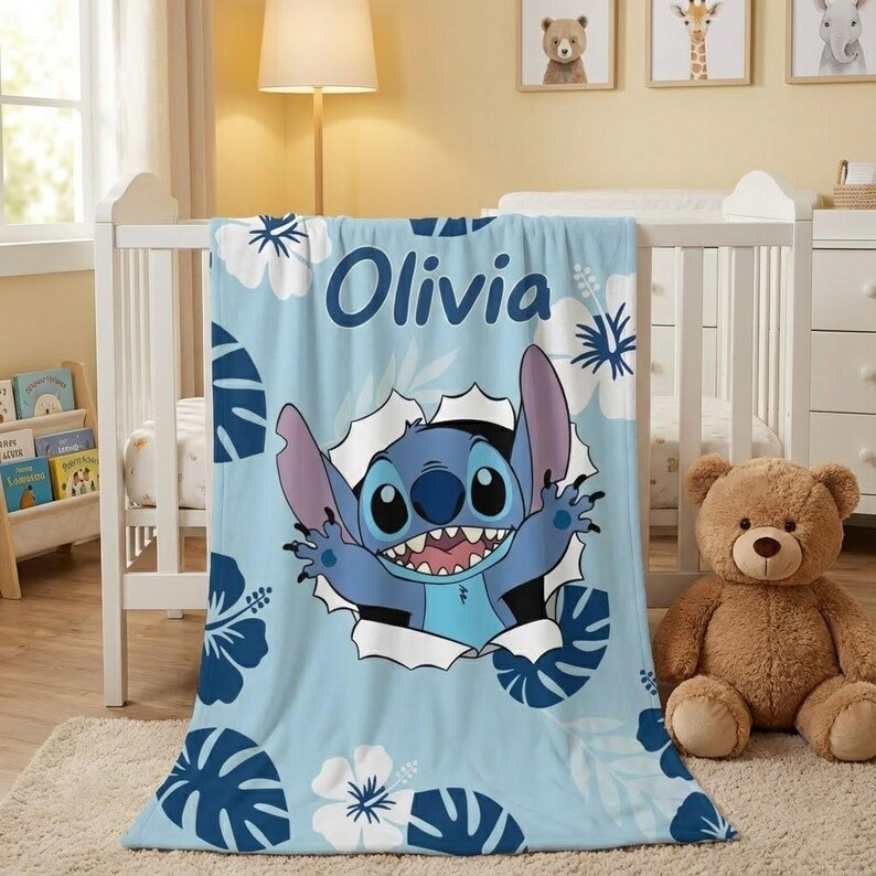 May include: A light blue baby blanket featuring the character Stitch from Lilo & Stitch. The blanket has a floral pattern with the name "Olivia" in dark blue. A brown teddy bear sits next to a white crib.