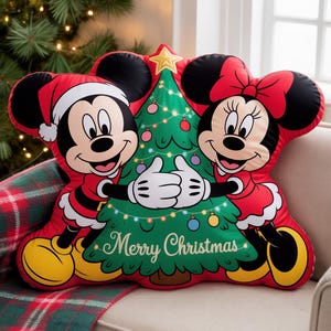 Peut inclure: Un coussin de Noël rouge en forme de Mickey et Minnie Mouse tenant un sapin de Noël décoré. Le coussin porte l'inscription "Merry Christmas" sur l'arbre. Mickey porte un bonnet de Père Noël et Minnie un nœud rouge.