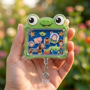 Op de afbeelding: Een groene badgehouder in de vorm van een kikker met een helder rechthoekig venster met diverse Toy Story-karakterstickers, waaronder Buzz Lightyear, Woody, Slinky Dog en de Aliens tegen een blauwe achtergrond met het Toy Story-logo.