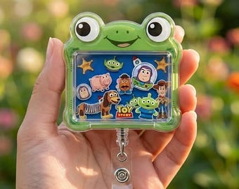 May include: A green frog-shaped badge reel featuring a blue background with Toy Story character stickers including Buzz Lightyear, Woody, Slinky Dog, Mr. Potato Head, Hamm, and the green aliens, with a retractable cord and metal clip.