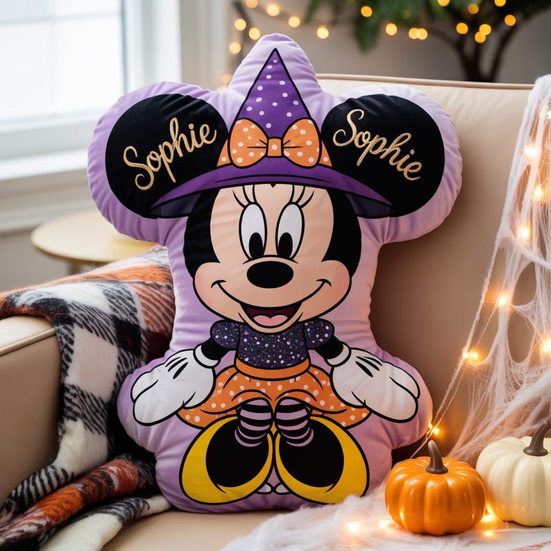 May include: A light purple shaped pillow featuring a cartoon mouse character wearing a purple witch hat with white polka dots and an orange bow. The name Sophie is embroidered in gold script on both black ears. The character wears a dark dress.