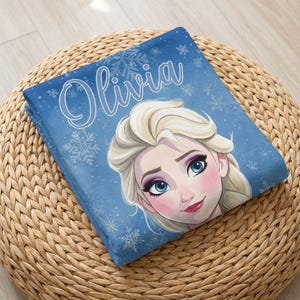 Pode incluir: Um cobertor azul dobrado com uma imagem de desenho animado de uma mulher com cabelo loiro e o nome "Olivia" em escrita branca. O cobertor tem um padr&atilde;o de floco de neve e est&aacute; sobre uma superf&iacute;cie tecida marrom.