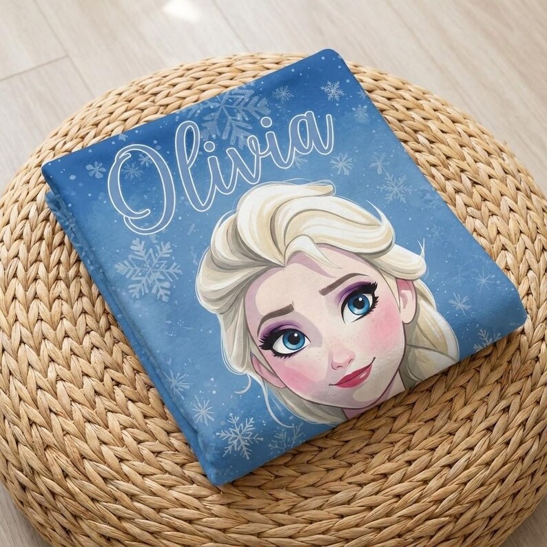 Pode incluir: Um cobertor azul dobrado com uma imagem de desenho animado de uma mulher com cabelo loiro e o nome "Olivia" em escrita branca. O cobertor tem um padr&atilde;o de floco de neve e est&aacute; sobre uma superf&iacute;cie tecida marrom.