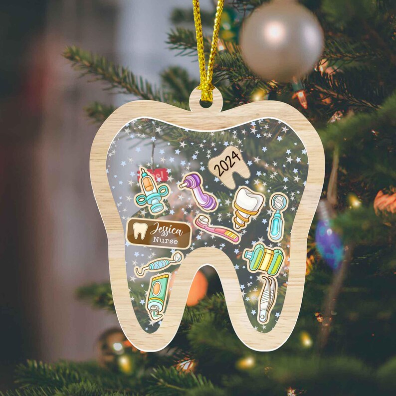 May include: A wooden Christmas ornament shaped like a tooth, featuring dental tools and the text "2024." The ornament includes the name "Jessica Nurse" and is hanging on a Christmas tree. The ornament has a gold string for hanging.
