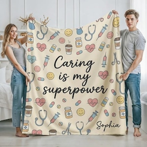 Puede incluir: Una manta beige con un patrón repetido de ilustraciones médicas, incluyendo estetoscopios, jeringas y pastillas. El texto "Caring is my superpower" está escrito en negro, con el nombre "Sophia" en la parte inferior.