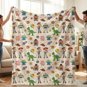 May include: A cream-colored blanket featuring a repeating pattern of cartoon characters, including a green dinosaur, a pig, a cowboy, and a space ranger. Other elements include a rocket, a ball, and a sheriff's badge.