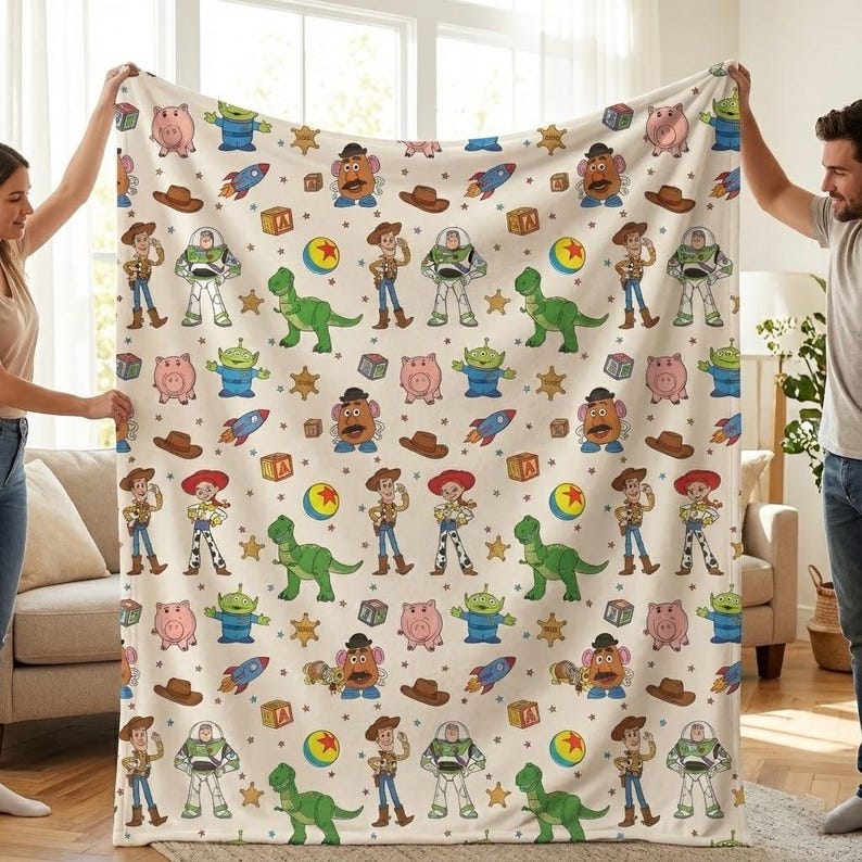 May include: A cream-colored blanket featuring a repeating pattern of cartoon characters, including a green dinosaur, a pig, a cowboy, and a space ranger. Other elements include a rocket, a ball, and a sheriff's badge.