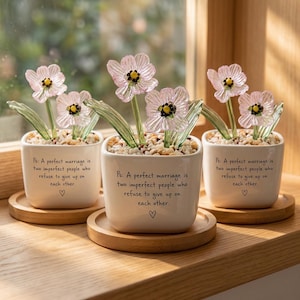 Puede incluir: Tres pequeños maceteros cuadrados de cerámica blanca, cada uno con un arreglo floral de cristal con pétalos rosas y hojas verdes. Las macetas están sobre posavasos de madera. El texto en las macetas dice: "Ps: Un matrimonio perfecto son dos personas imperfectas que se niegan a rendirse."