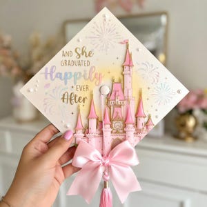 May include: A diamond-shaped graduation cap topper featuring a pink castle illustration, fireworks, and the text "And She Graduated Happily Ever After" in gold and pastel lettering. The topper is decorated with white pearls and a pink satin bow.