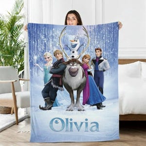 Puede incluir: Una manta azul con personajes de la pel&iacute;cula animada Frozen, incluyendo a Elsa, Anna, Kristoff, Olaf y Sven. La manta tiene un fondo de bosque invernal y el nombre "Olivia" en fuente azul claro.