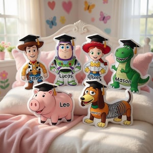May include: A collection of plush character pillows featuring graduation caps. Characters include Woody, Buzz Lightyear, Jessie, Rex, a pig, and a dachshund, each with a name. The pillows are on a bed with a pink blanket.