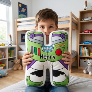 May include: A white, green, and purple letter "H" shaped pillow featuring a cartoon design. The design includes the name "Henry" and a space ranger emblem. The pillow is in a child's bedroom.