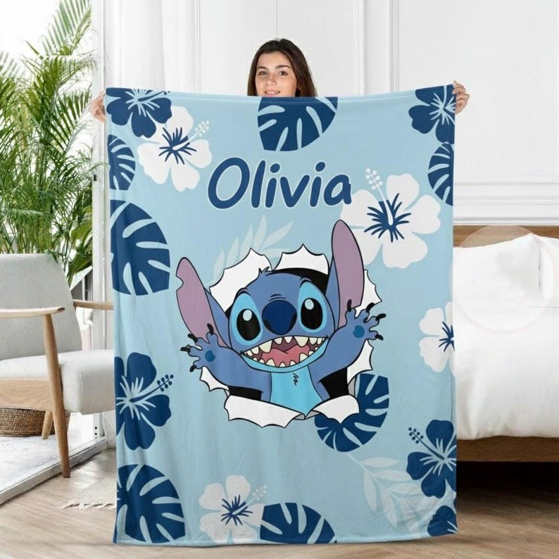 May include: A light blue blanket featuring a cartoon character, surrounded by white and dark blue floral and leaf patterns. The name "Olivia" is printed in blue script. The character is breaking through a white hole.